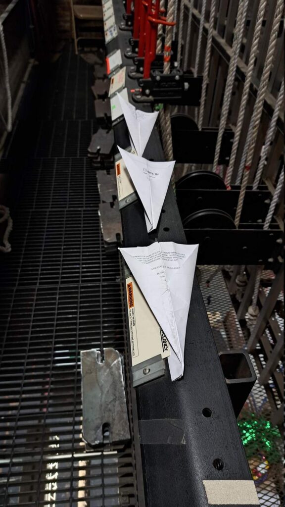 Three folded paper airplanes lined up on a backstage fly rail among ropes and metal hardware at Tupelo Community Theatre.