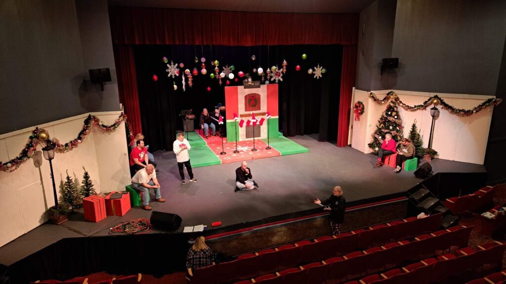 Wide shot of the Lyric Theatre stage with actors spaced across the Christmas set while the director gives notes from the front row.