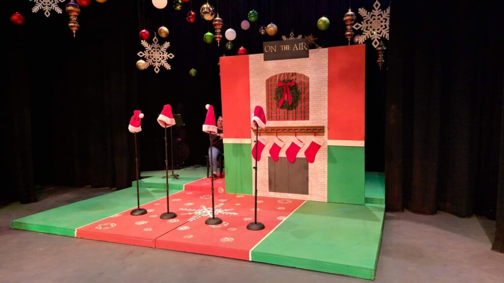 Close shot of red-and-green stage with snowflake floor, fireplace backdrop, and microphones each wearing a bright red Santa hat.