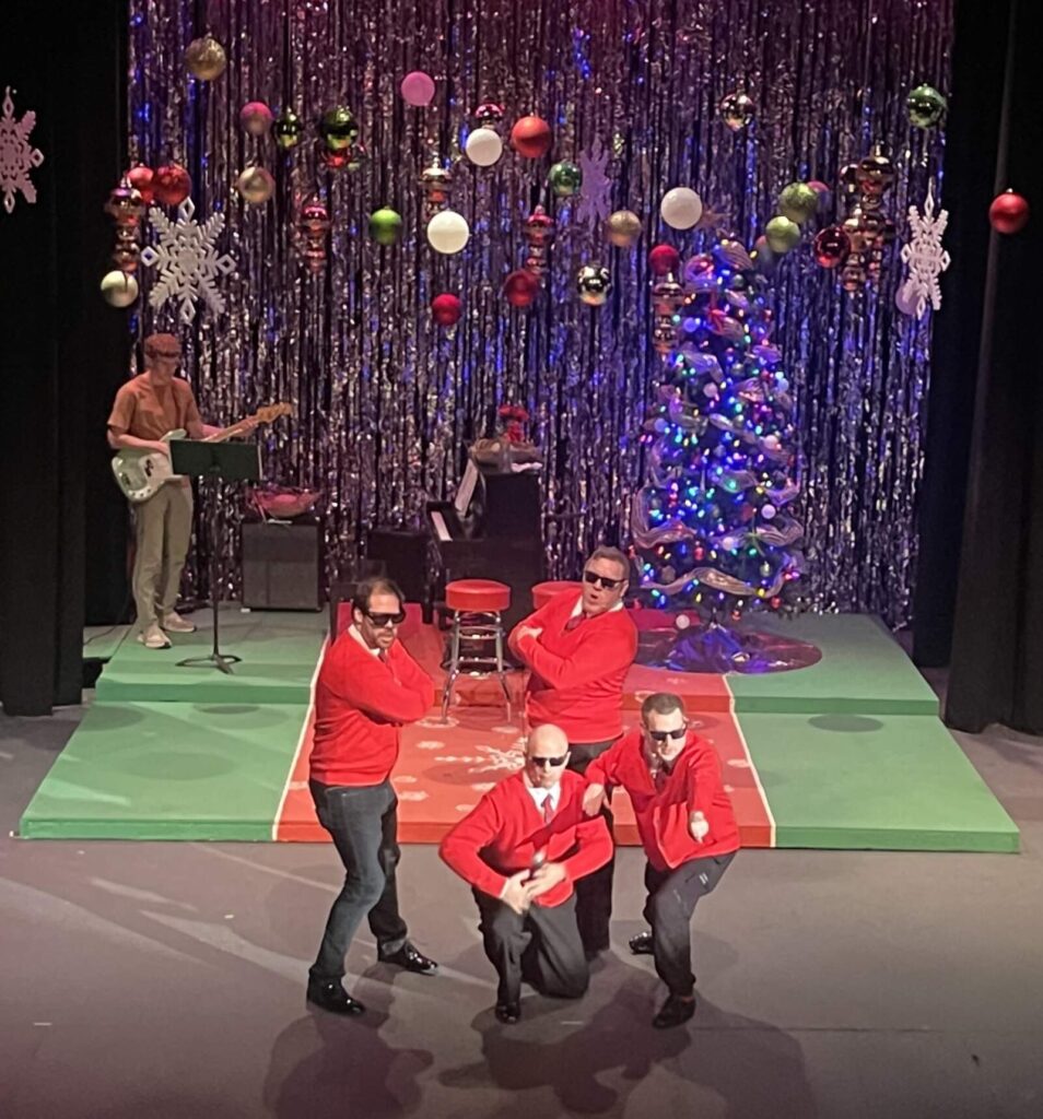 Group of four male performers in red sweaters, black pants, and sunglasses posing in formation on a brightly lit holiday stage with silver tinsel backdrop.
