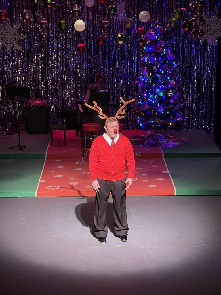 Man in a red sweater with reindeer antlers and a red clown nose stands alone in a spotlight on a Christmas-decorated stage.