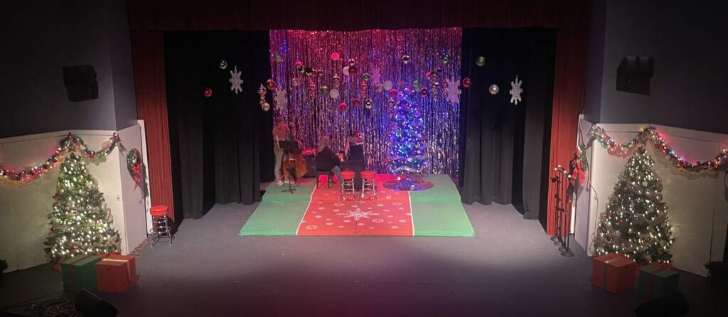 Full-stage photo showing musicians rehearsing on a central platform framed by Christmas trees, garlands, and a shiny backdrop of hanging baubles.