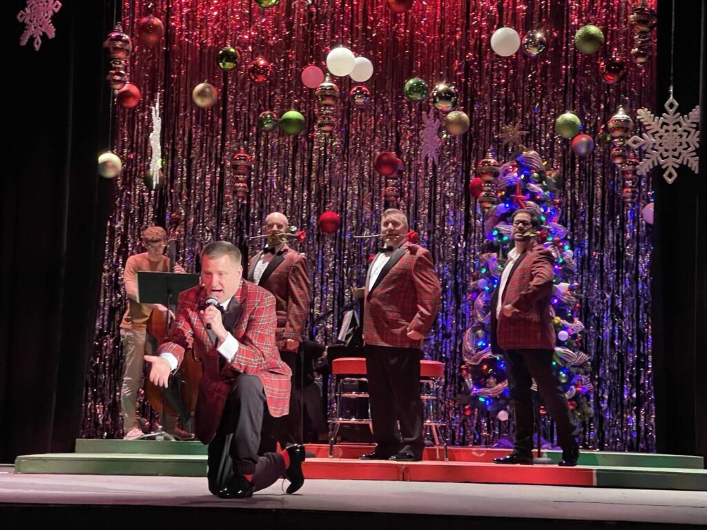 Male singer in a red plaid jacket kneels at the front of the stage with microphone while three fellow performers and musicians stand behind him on a decorated holiday backdrop.
