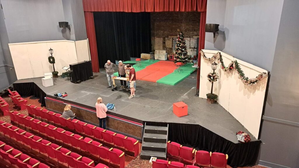 Wide view of the Plaid Tidings stage with red and green platforms, Christmas tree, and crew gathered at a rehearsal table.