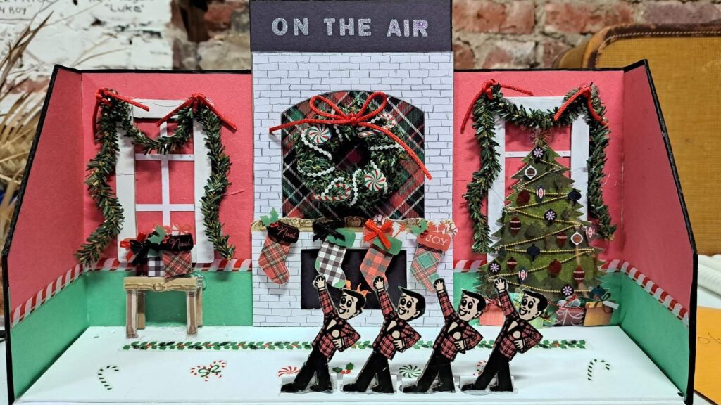 Miniature Christmas living room with “On The Air” sign, fireplace, stockings, garlands, tree, and tiny Plaid figures in front.
