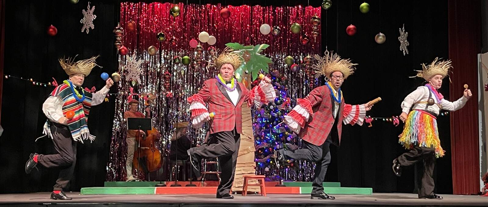 Four male performers in plaid jackets, straw hats, and tropical accessories dance with maracas in front of a Christmas tree and tinsel backdrop.