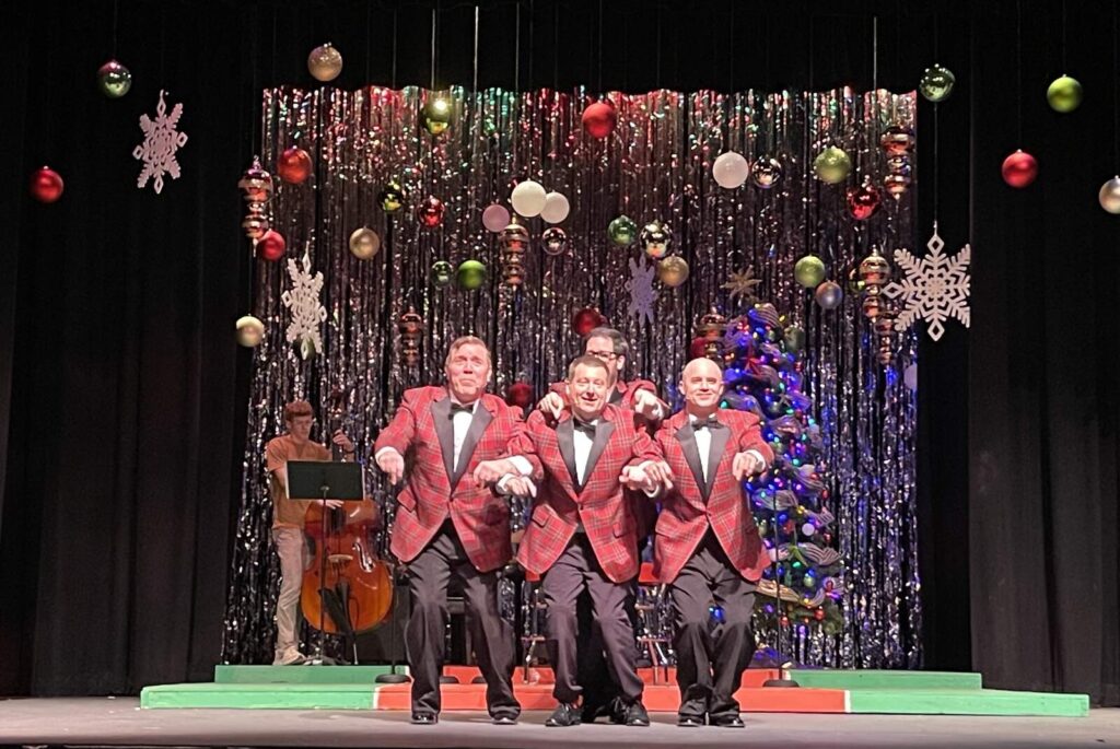 Group of four performers in matching plaid tuxedo jackets move in unison with hands held out as part of a choreographed number on a tinsel backdrop.